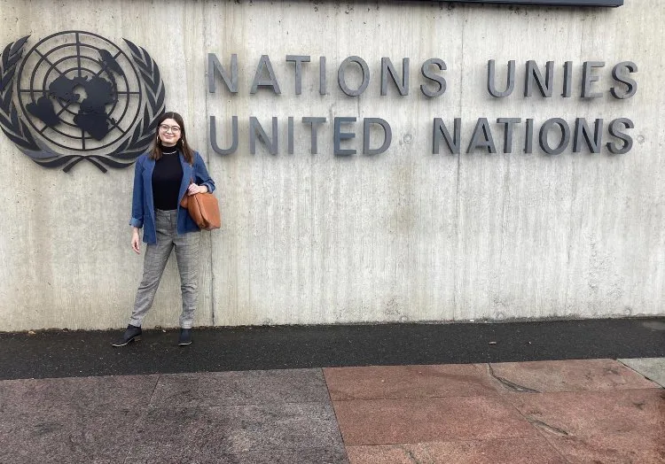 Lela Tolajian, a woman with a light skin tone and short brown hair, poses in front of a sign that reads, “Nations Unies United Nations.” A globe symbol is on the left. 