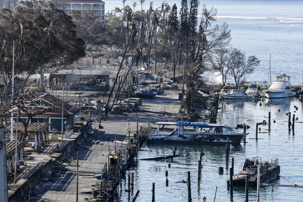 Destruction from wildfires seen on a Watercraft near downtown Lāhainā. Burned structures and vehicles are in the background. Several boats sit on the water. 