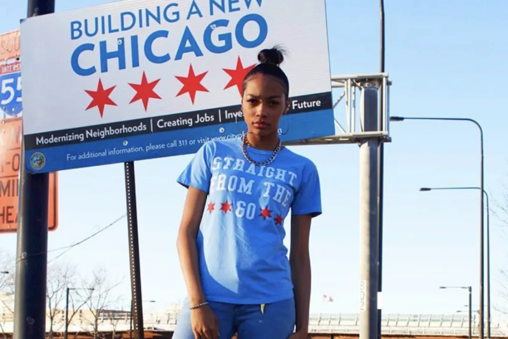 Raven Smith stands in front of a construction sign with the Chicago flag that reads, “Building a new Chicago.” She stares at the camera and is wearing a blue shirt that reads, “Straight from the GO.” Her hair is in a bun.