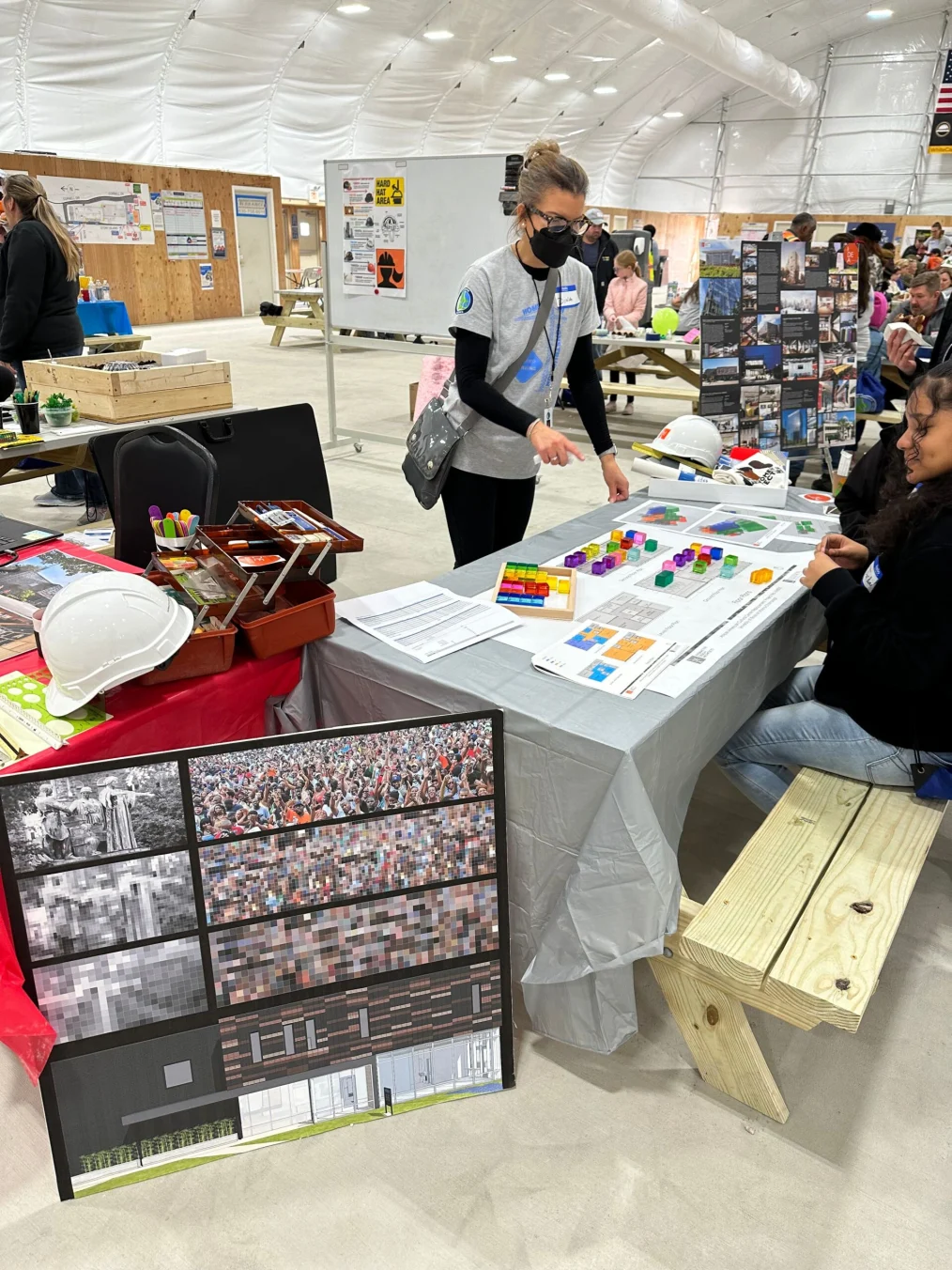 Dina Griffin stands behind a table at the Lakeside Alliance “Who Builds the World? GIRLS!” event. She is wearing a mask, glasses, and gray t-shirt as she explains a diagram to a young woman sitting at the table. On the table are rainbow colored cubes and architectural designs. 