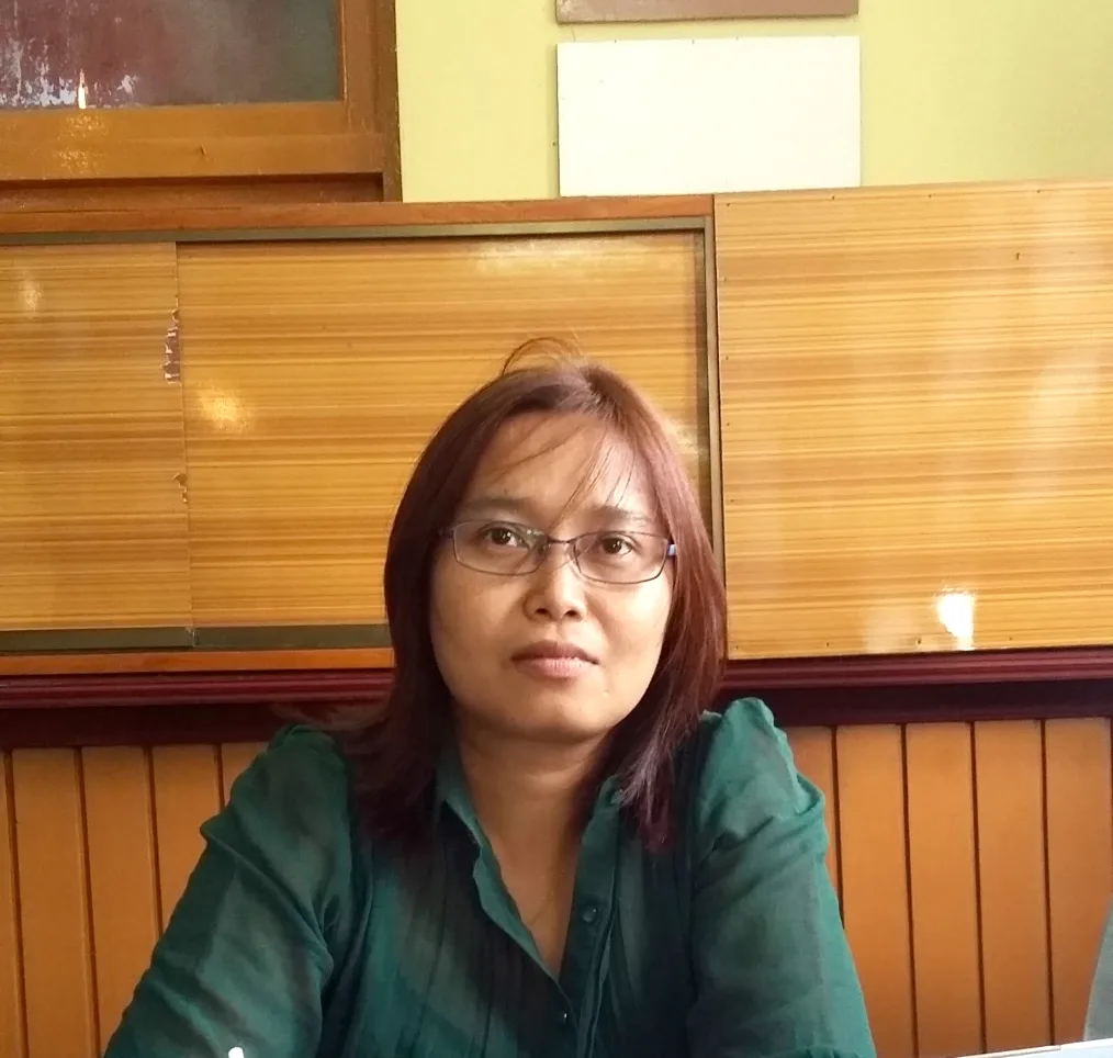A woman with a neutral light skin tone sitting down at a booth wearing glasses and a dark green formal shirt and burgundy shoulder length hair 