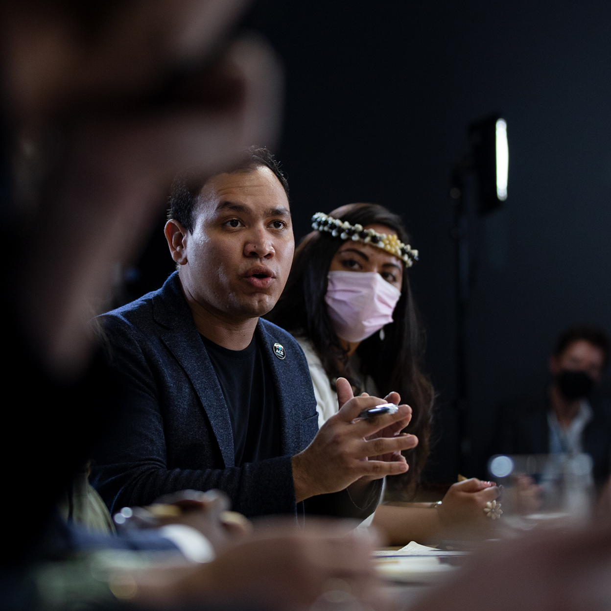 Juan Carlos Monterrey Gómez, a man with a medium skin tone, speaks from a conference table. He is wearing a navy blazer and black shirt. A woman with a light skin tone is wearing a pink face mask next to him. A blurred person is in the foreground. 