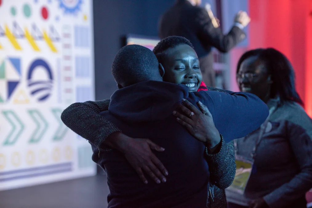 Two people hug each other. The photo is dimly lit, but other people can be seen out-of-focus in the background.