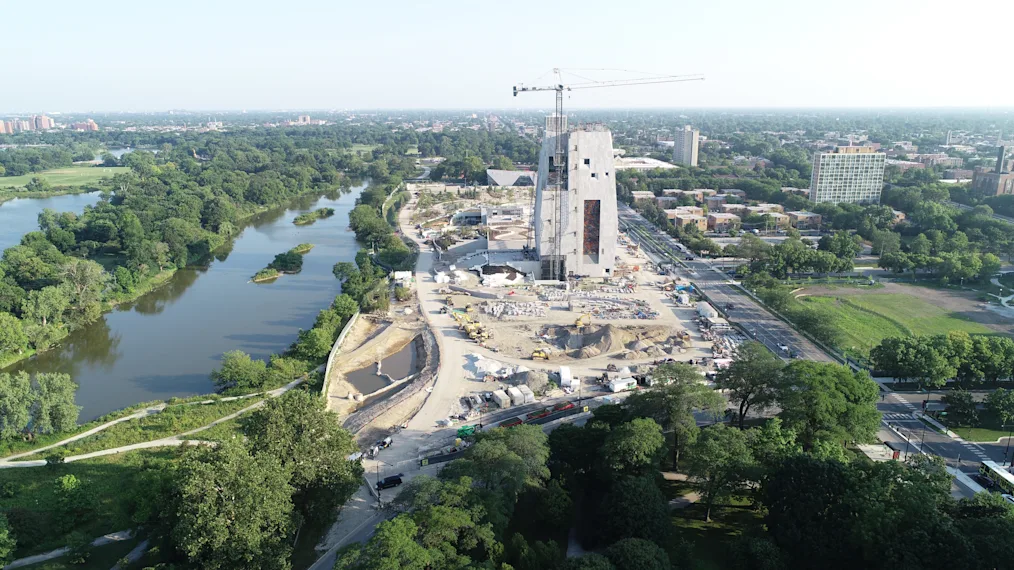 An image of the Obama Presidential Center campus with construction. The picture features an aerial view of the entire campus. In the center is the Museum Building with a crane on top. To the left of the Museum Building is a lake and trees. To the right of the building is Jackson Park.