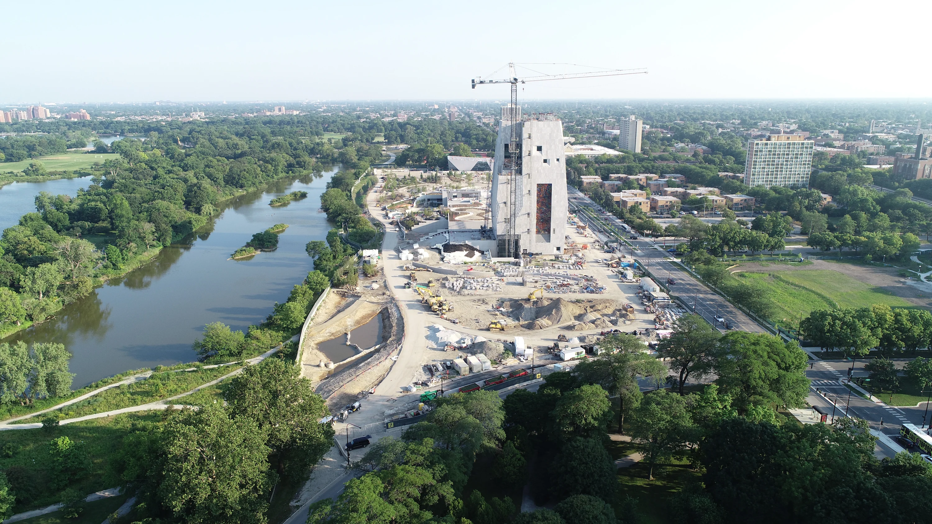 An image of the Obama Presidential Center campus with construction. The picture features an aerial view of the entire campus. In the center is the Museum Building with a crane on top. To the left of the Museum Building is a lake and trees. To the right of the building is Jackson Park.
