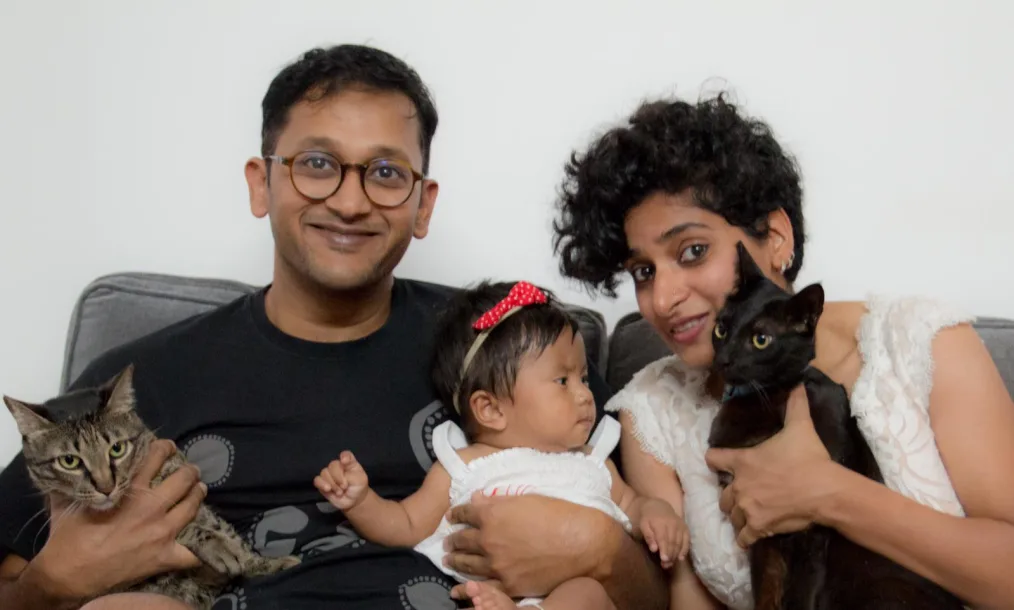 A man, a baby and a woman sit on a gray couch. The man holds a tabby cat and the baby, while the woman holds a black cat.