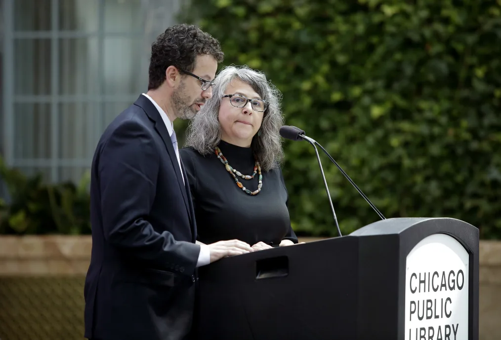 

An older woman with glasses and grey hair and a light skin tone stands next to an older man with black hair, glasses, and a light skin tone over a podium that reads "Chicago Public Library"