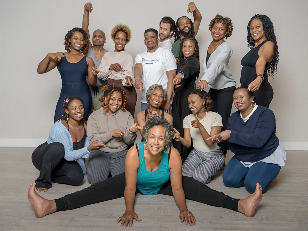 A group shot of Julia Perkins, a woman with a light medium skin tone, holds a split at the front of a group of people with a range of light to dark skin tones smiling and posing for a photo. 