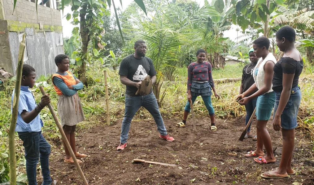  Obama Leader Njeke Joshua Egbe, a man with a dark skin tone and low black hair, teaches a farming course to six girls in his community. The girls have dark skin tones and are formed in a semi circle. 