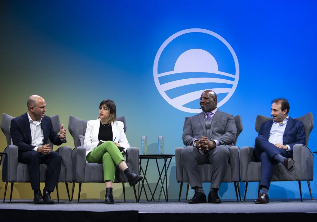 A group of four people sitting down on gray chairs with small microphones  
connected to their ears, appear to be engaging in a meeting together. The
 Obama Foundation logo appears on a blue background in white text.