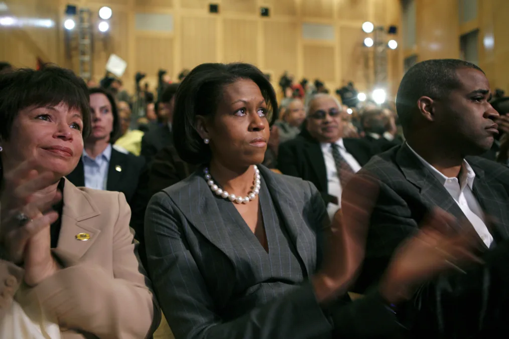 Michelle Obama, her eyes watering with tears and her mouth pursed, sits in a room full of people. Bright spotlights are on the back wall. Everyone looks dressed in formal suit attire.