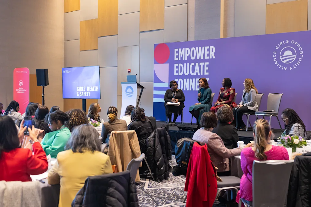 A group of women with a range of light to dark skin tones sit around tables in a room. In the background, a purple banner reads, “Empower, Educate, Connect.”