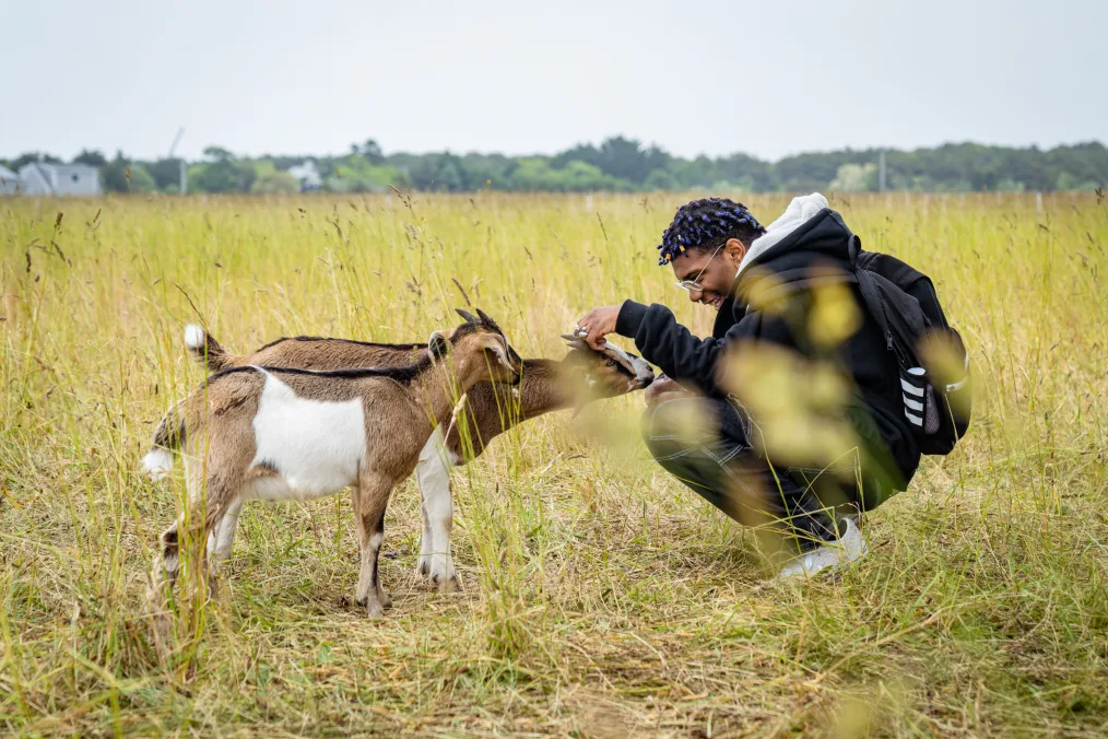 A young man with a deep skin tone smiles as he crouches in front of two brown and white goats. They are in an open field. The young man is wearing all black, a backpack, and glasses.