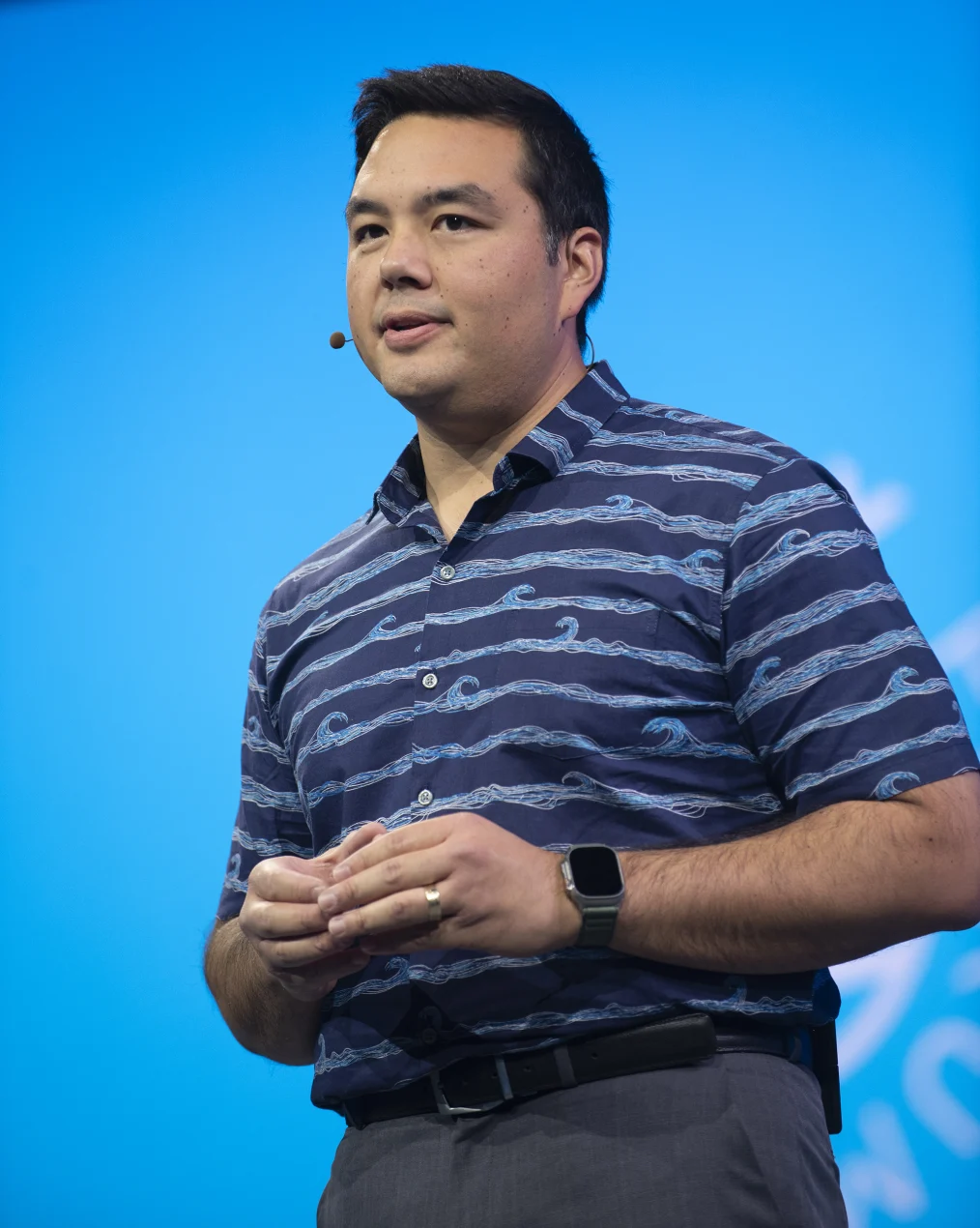 A middle aged man with light skin with warm under under tones, short slick black hair stands giving a speech. He has a mic attached coming behind his right ear. He has a polo shirt with waves, an apple watch on his left wrist, a black belt on, and grey pants. The background is a blurred light blue wall. 