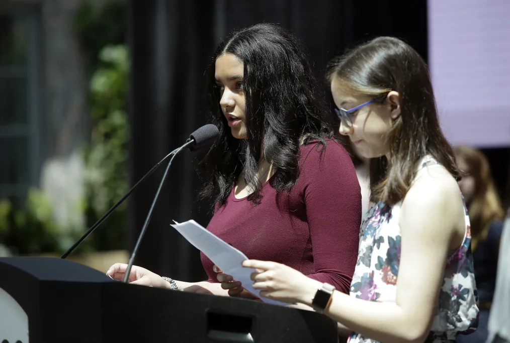 A woman with medium skin tone and black hair speaks at a podium next to a woman with a light skin tone, glasses, and brown hair