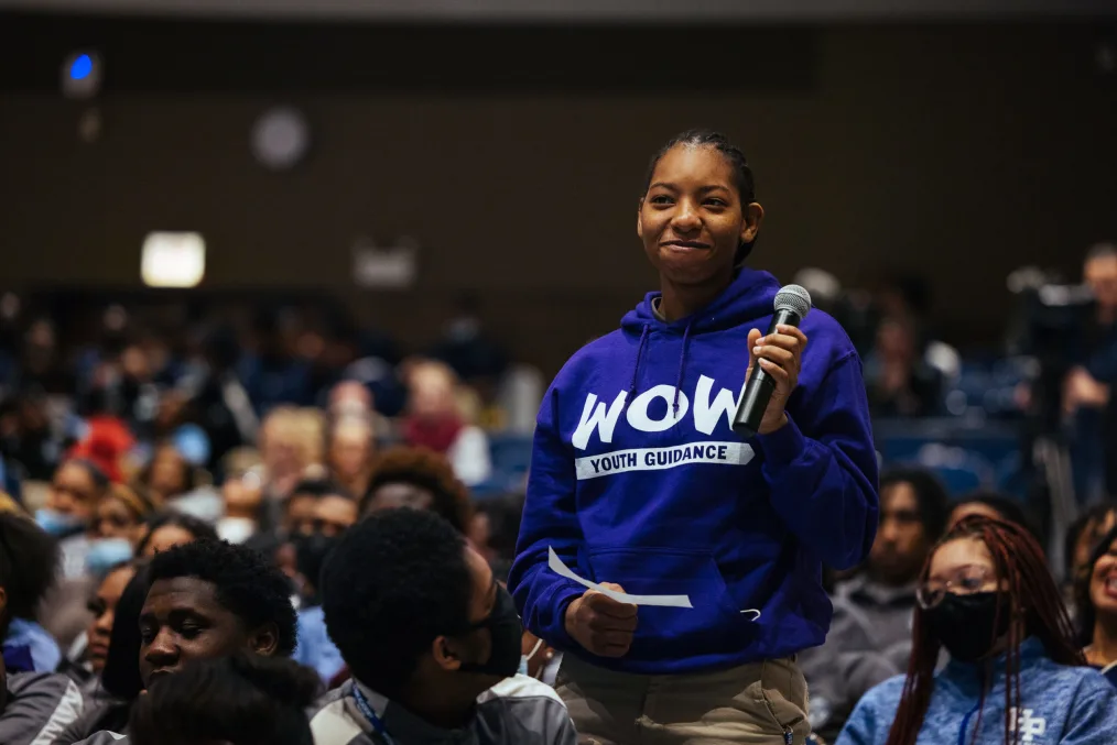 A young girl, who is medium-deep toned, stands to say something during an event. She has cornrows, a nervous smile, and is wearing a blue shirt that says boldly "WOW, Youth Guidance"