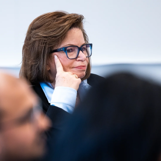 Valerie Jarrett sits listening with her hand up to her face. She wears blue and black glasses, a light blue blouse and a black jacket.