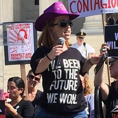 Rebecca holds a microphone as she speaks at a rally at the Supreme Court. She is a red headed woman with a light skin tone. She is wearing a "I've been to the future, we won'' shirt and a purple velvet cowboy hat with a gold uterus in the middle. People hold signs in the background. 