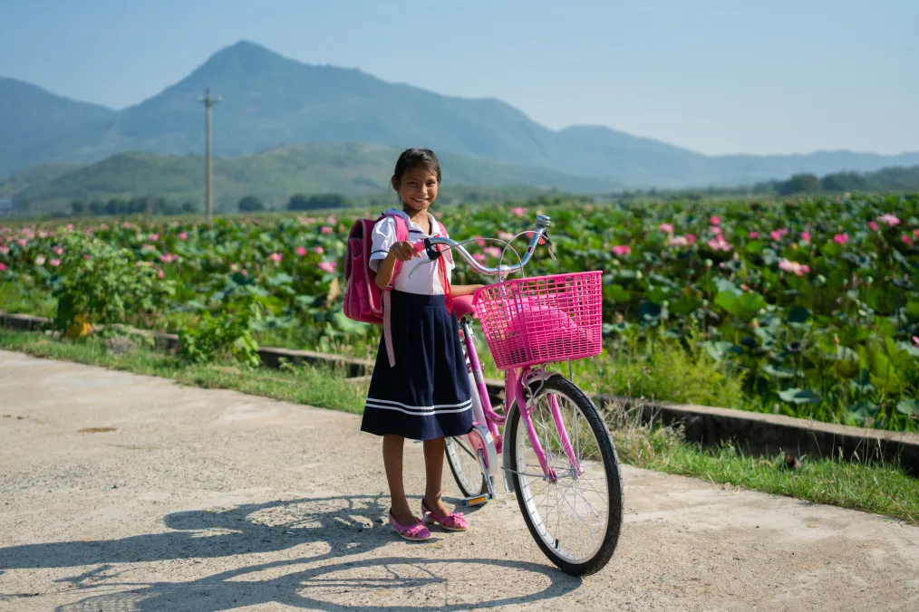 A young girl with a warm olive tone stands in front of a field with azelas in the background, wearing a white collared shirt, navy blue skirt with two white stripes at the bottom, a pink backpack, pink shoes, while holding the handles of a pink and white bike with a pink basket on it.