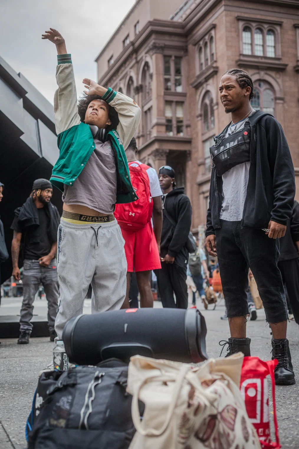 The image is a photo of several men on a street outside. Behind the men is a brown stone building. To the left of the men is an abstract statue. There are several men standing in a circle in the foreground of the image. In the front of the image are two men. The first man is standing in a position to symbolize throwing a basketball shot. He has a light deep medium complexion, a large black afro and is wearing a green and beige lettermans jacket, a grey shirt, and grey sweatpants. The second man has a medium deep complexion, a goatee, and cornrows in his hair. He is wearing a black hoodie, a grey shirt, cut off black pants cut to the calf, and black combat boots. He has a fanny pack sitting on his neck. In front of the men are several backpacks sitting on the ground.