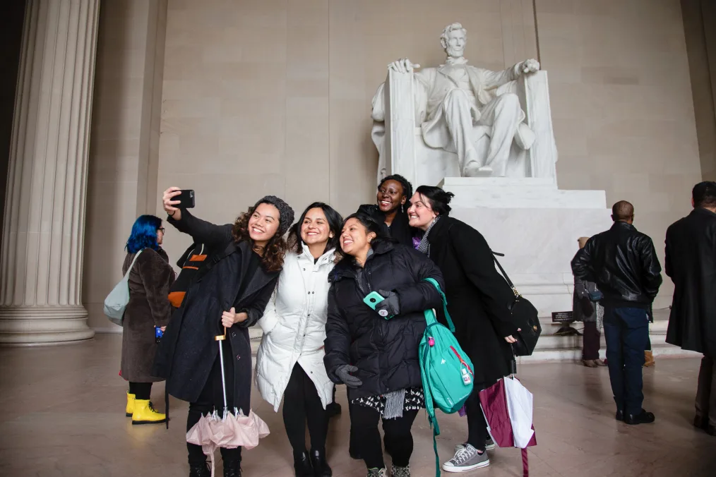 A group of people pose for a selfie in front of the Lincoln Memorial. They wear warm winter coats and carry backpacks and umbrellas.