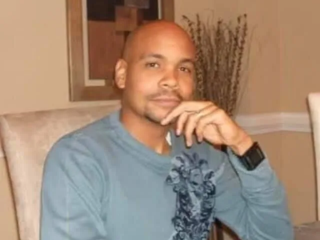 Roderic Walker, a Black man with a medium skin tone, stares into the camera as his hand rests on his cheek. He has no hair and is wearing a gray sleeved shirt.