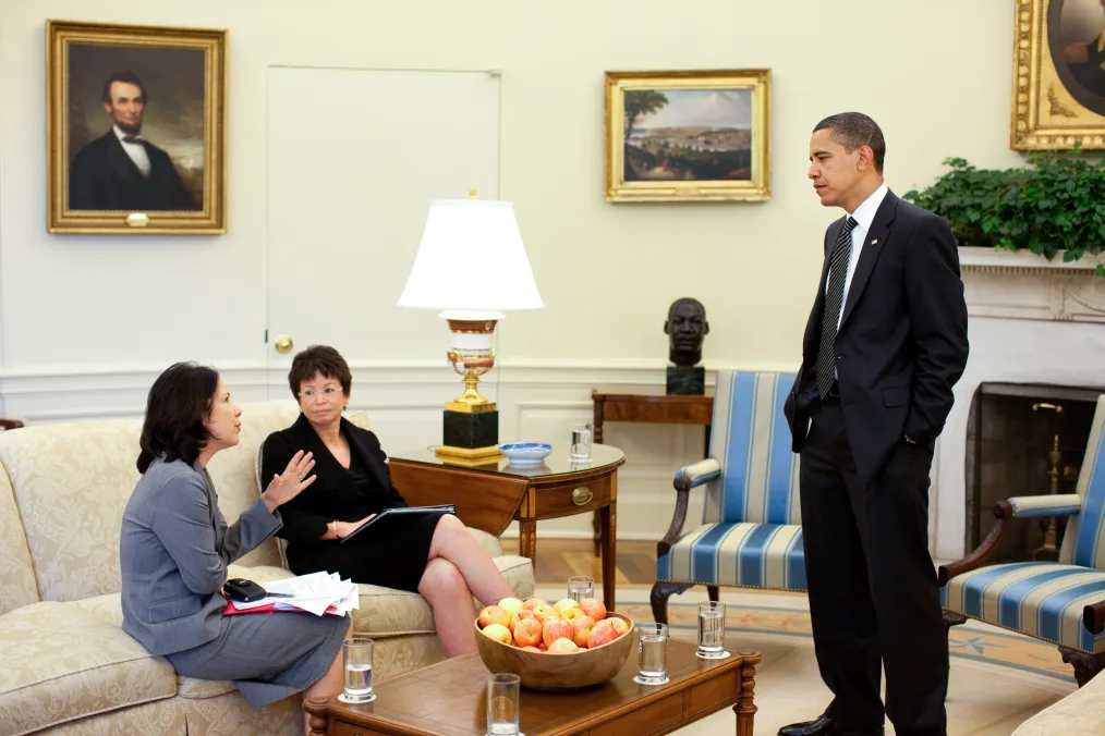 Nancy-Ann DeParle, a woman with a light skin tone and dark hair, sits on a couch next to Valerie Jarrett, a Black woman with a light skin tone as she speaks to President Obama. President Obama is standing. He is wearing a black suit. Nancy-Ann DeParl and Valerie Jarrett are wearing gray and black skirt suits.