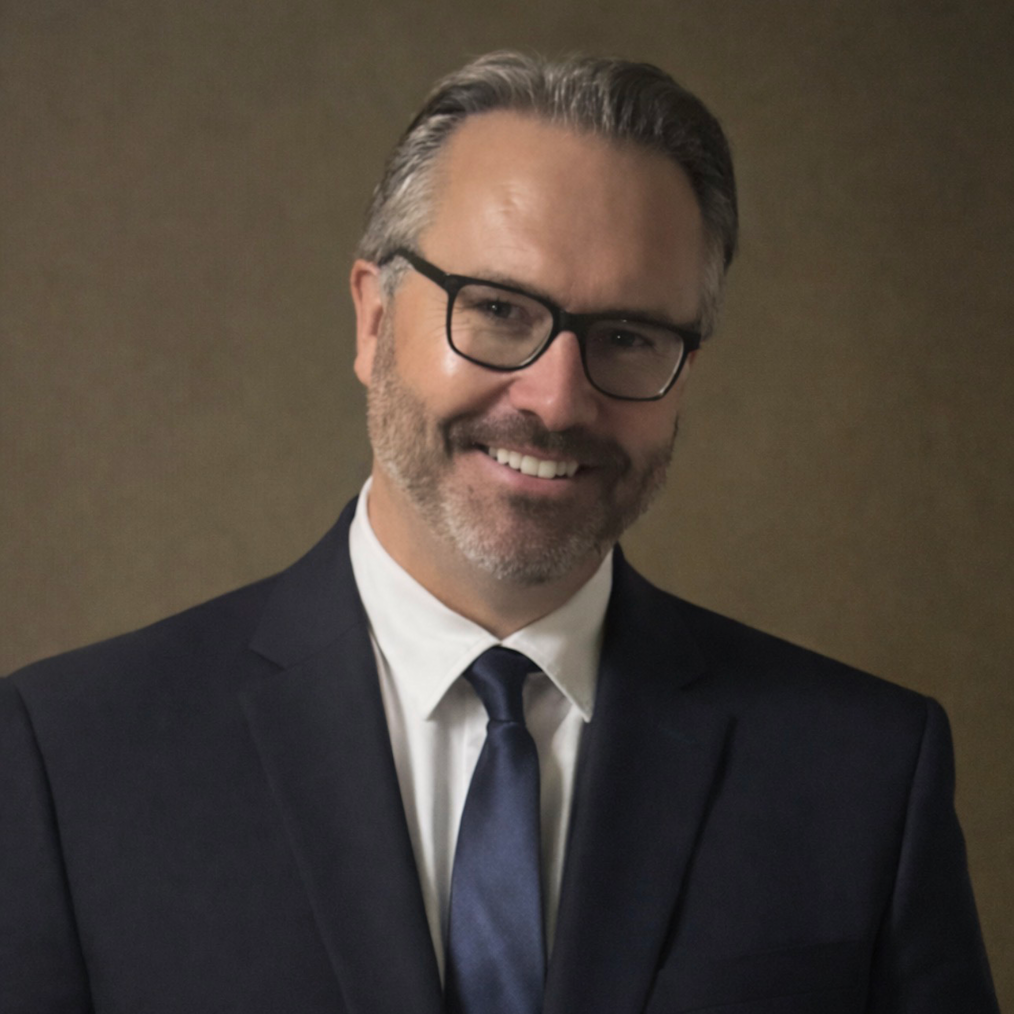 A headshot of a white man with gray hair, glasses, blue tie and blue suit jackets. His arms are crossed and he smiles slightly with his teeth showing.