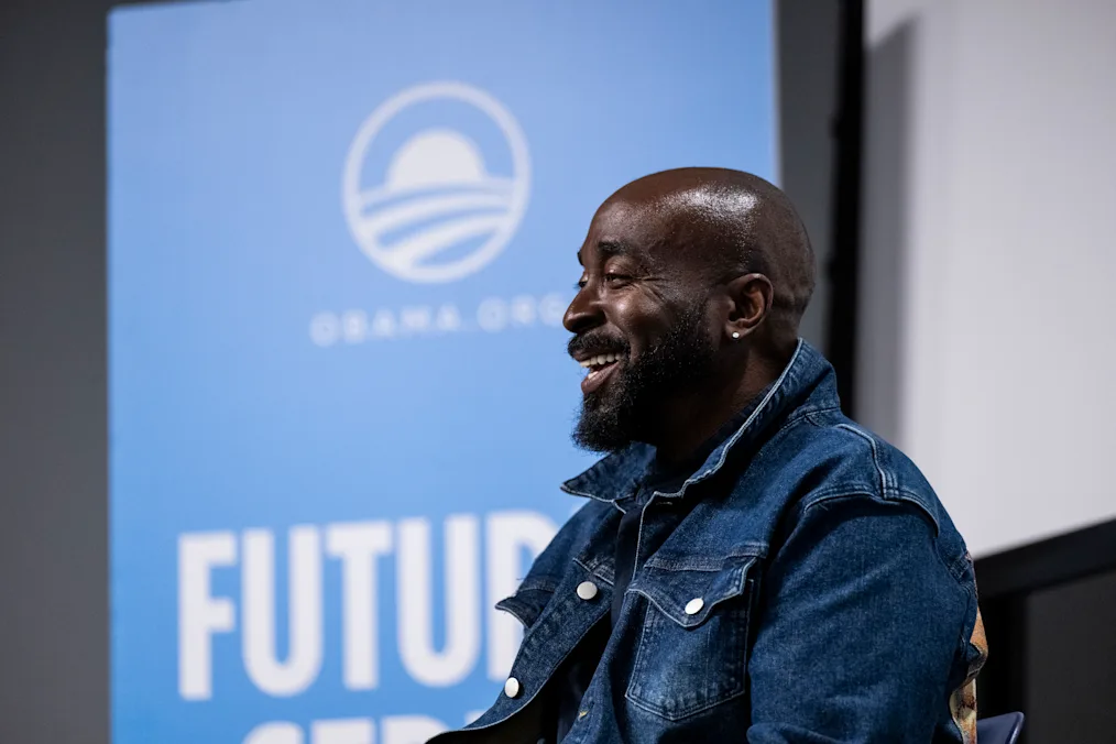A side profile of Eric Williams, a Black man with a dark skin tone and no hair, smiling at an audience. He is wearing a denim shirt with a flipped collar. A blurred blue banner in the background reads, “Obama.org.”