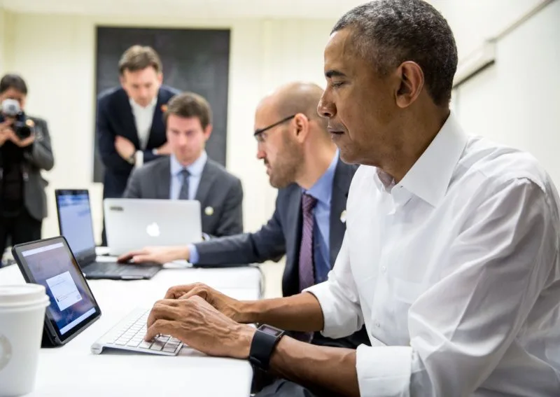 Barack Obama sits at a table typing at a keyboard. Several other men do the same, while a man and a person with a camera look on.