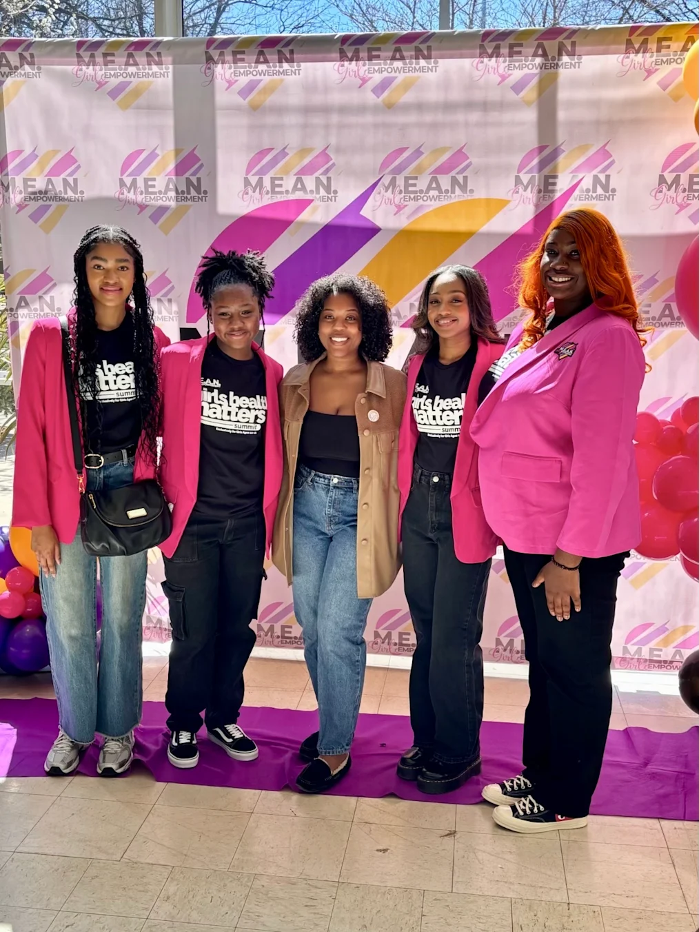 A group of five Black women and girls pose in front of a M.E.A.N. Girls Empowerment branded step-and-repeat. Four are wearing pink blazers and black shirts that read “Girls Health Matters.”