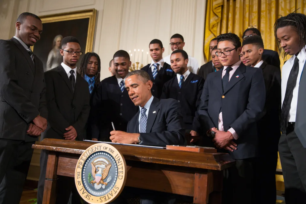 A group of young men wearing suits stand behind lightly smiling as they watch President Obama sign a document at a brown table with a presedential seal. The setting is a regal office or room.  