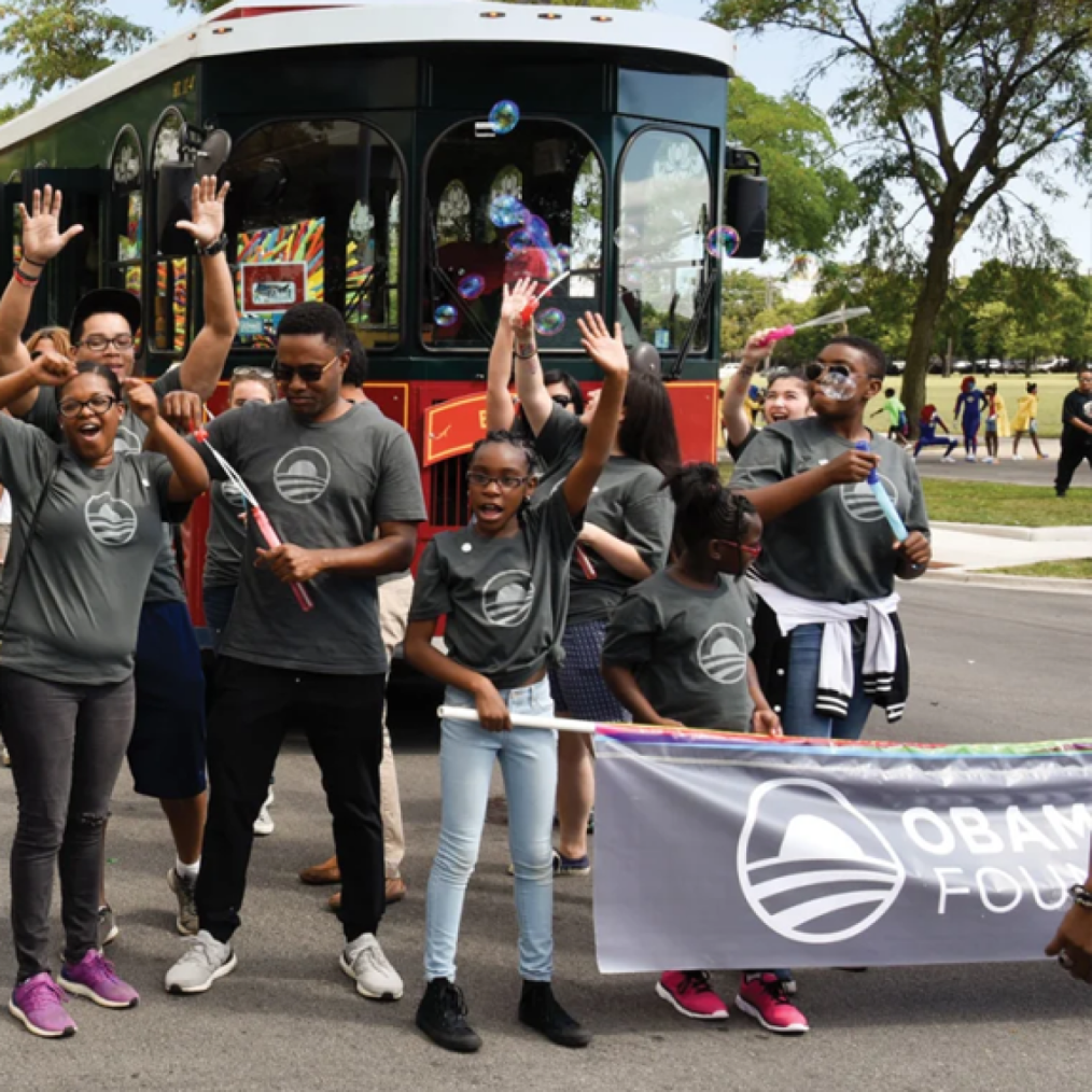 A group of people with a range of light to deep skin tones walk in front of a trolley. They are all wearing gray shirts with the Obama Foundation rising sun logo and holding up Obama Foundation signage.