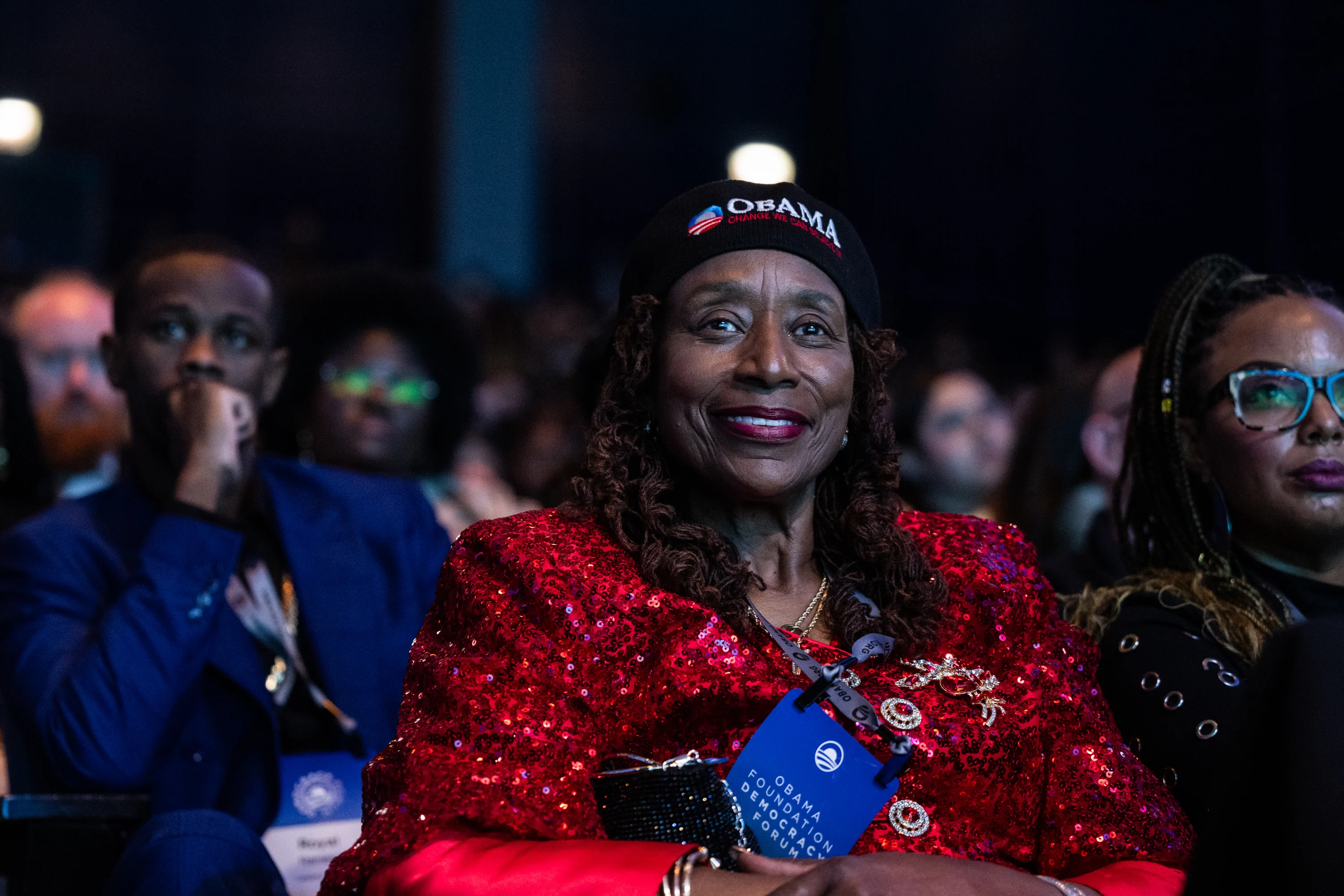 A dark-skinned woman smiling sits in an audience wearing a sparkling red sequined blazer and a black beanie with "Obama" embroidered on it. 