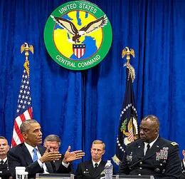 President Barack Obama, wearing a dark blue suit and tie, sits next to a man with a deep skin tone wearing a black uniform jacket with medals and patches. There are four people of a light skin tone in the background wearing black uniform jackets as well. Everyone is sitting in front of a royal blue curtain with the United States Central Command symbol hanging in front of it, as well as two flags. 