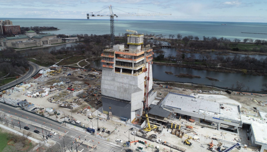 An aerial view of the South West corner of the Museum Building, the Forum, and part of the Library Building rooftops at the Obama Presidential Center. Construction materials can be seen covering the landscape.