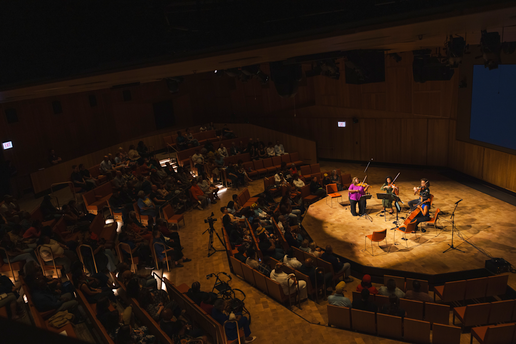 Four musicians perform on a sunlit stage before a seated audience in a multi-level, wood-paneled auditorium. Empty orange chairs and music stands surround the quartet on stage. Surrounding the stage area, audience members of diverse skin tones are seated in rows of wooden chairs arranged across step-down seating levels.

