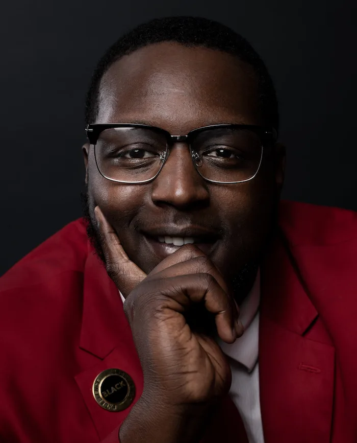 A portrait photo of a man with a deep skin tone wearing black frameless glasses, a white button-up shirt, and a red blazer, smiles with his hand on the left-hand side of the photo on his face in front of a black gradient background. 