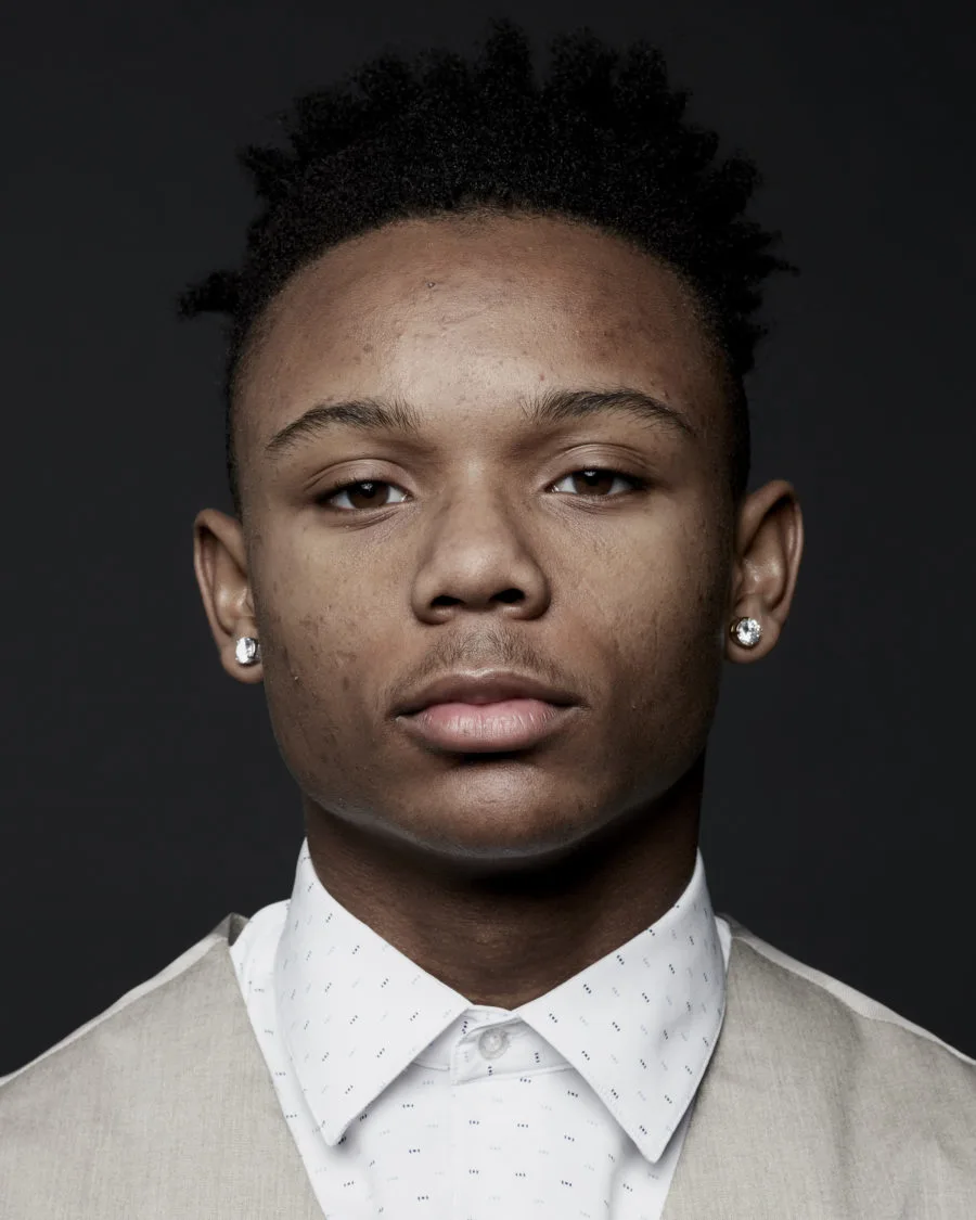 A portrait photo of a young man with a medium skin tone wearing a white button-up shirt and beige vest standing in front of a gradient black background.  