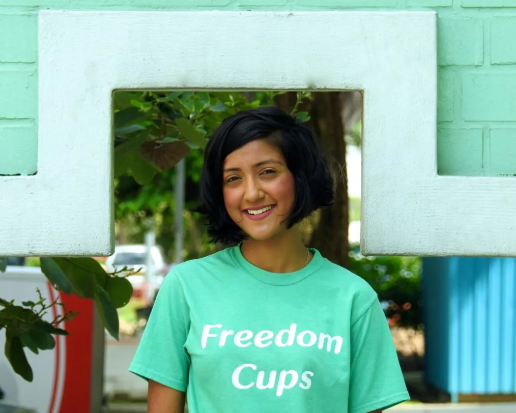 A woman with a medium deep skin tone smiles as she stands outside under a mint green structure. She has neck length black hair and is wearing a mint shirt that reads, "Freedom Cups."
