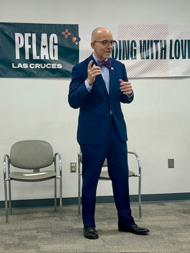 An image of Brian Bond giving a speech with PFLAG. Brian is standing in a conference room with a microphone in his hand and speaking.
Brian has a light olive complexion and a bald head. He is wearing glasses, a blue suit, a blue shirt, and a purple tie. Behind him is a white wall with a long horizontal poster on the wall. The first poster is a PFLAG Las Cruces logo poster with a dark blue background on one end. The other end of the poster is pink and white and says, "...with love."