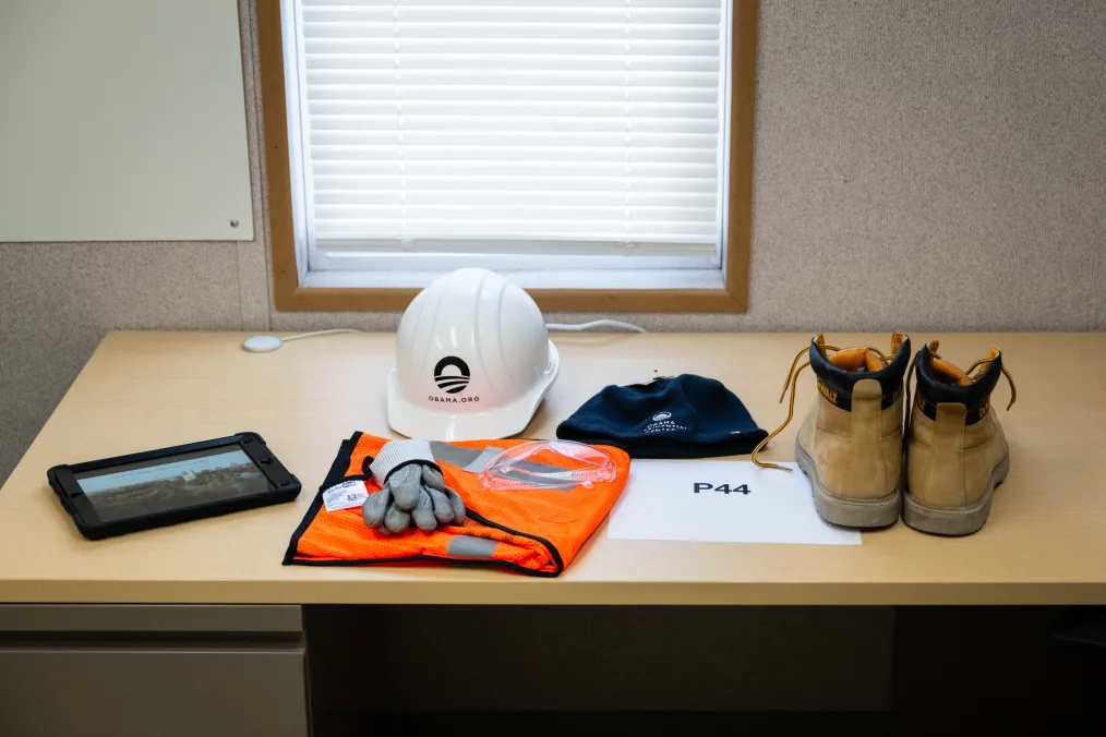 A black tablet, folded orange safety vest, gloves, a white hart hat with the Obama Foundation rising sun logo, a navy beanie with the rising sun logo, a sign that reads, “P44,” and chestnut work boots sit on top of a table at the site of the Obama Presidential Center construction site. 