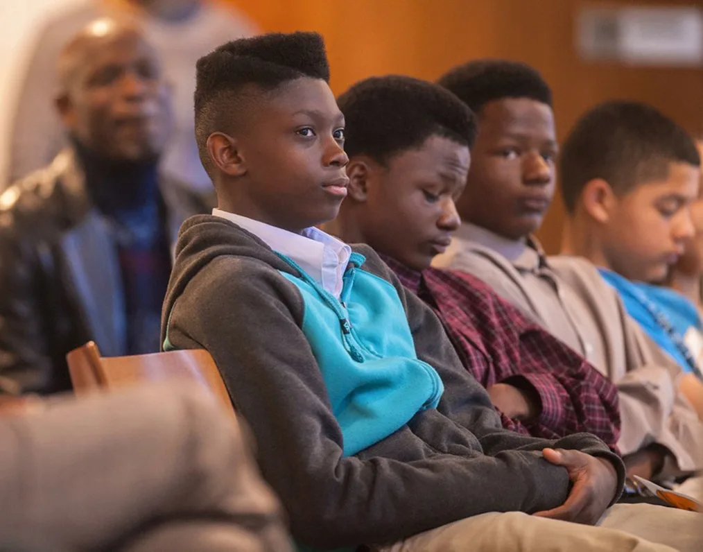 Several Black boys with engaged expressions sit with their hands in their lap, gazing to the right. 