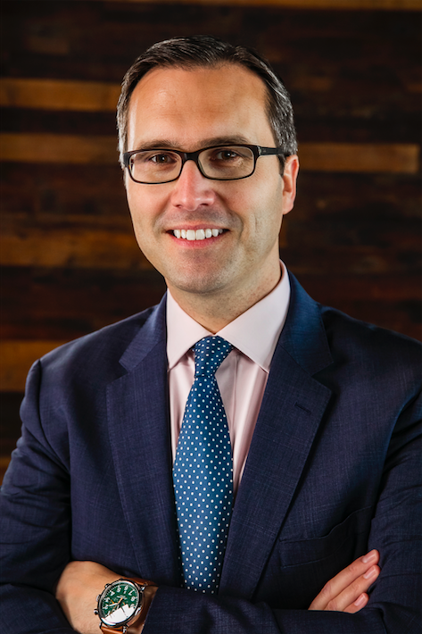 A headshot of a white man with gray hair, glasses, blue tie and blue suit jackets. His arms are crossed and he smiles slightly with his teeth showing.