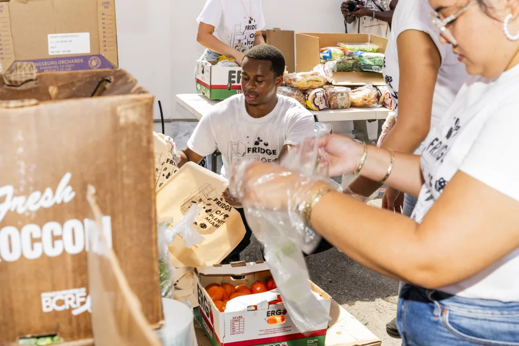 Jonathan Jean Charles, a Black man with a medium dark skin tone squats as he prepares produce bags that read, “Fridge of Plenty Miami.” He is surrounded by three others with medium skin tones who are out of frame putting together bags. Vegetables are on the tables. 