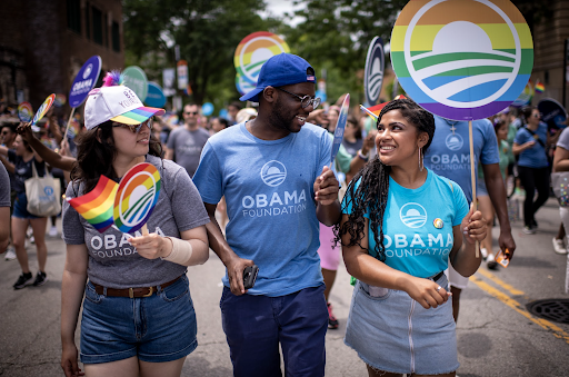 Two women with light medium skin tones walk in between a Black man with a deep skin tone. He is wearing glasses and a hat. All three are wearing Obama Foundation T-shirt’s and holding Pride Month flags. In the background is more signs and people.