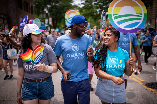 Two women with light medium skin tones walk in between a Black man with a deep skin tone. He is wearing glasses and a hat. All three are wearing Obama Foundation T-shirt’s and holding Pride Month flags. In the background is more signs and people.