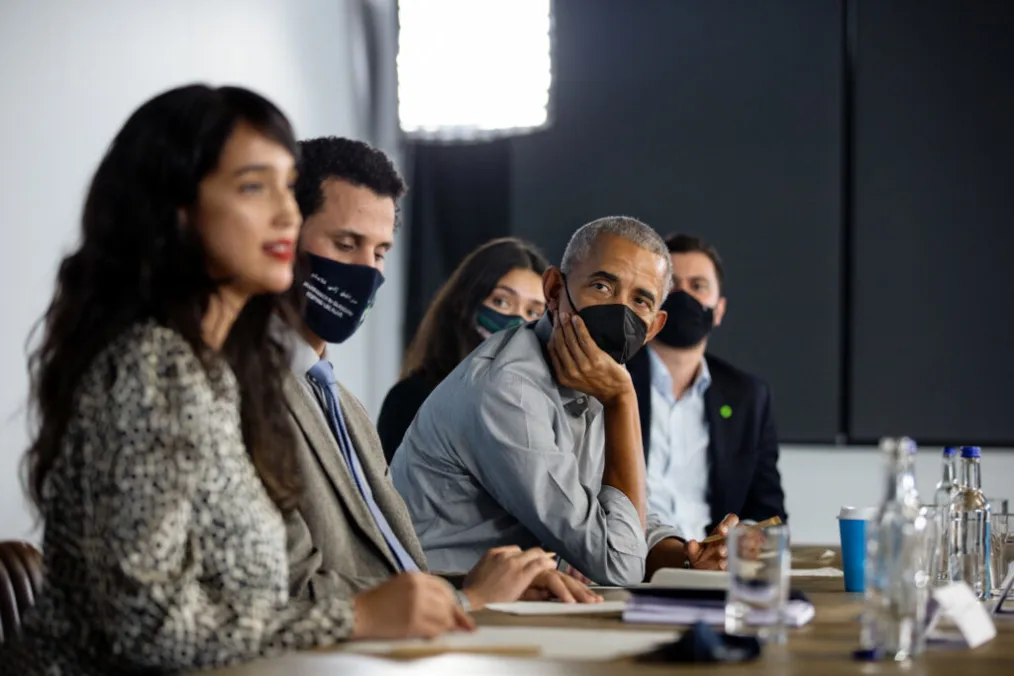 President Obama leans forward with his hand upon his chin, looking at a dark haired woman who is gesturing to the room. President Obama wears a black mask, and other masked people sit on either side of him.