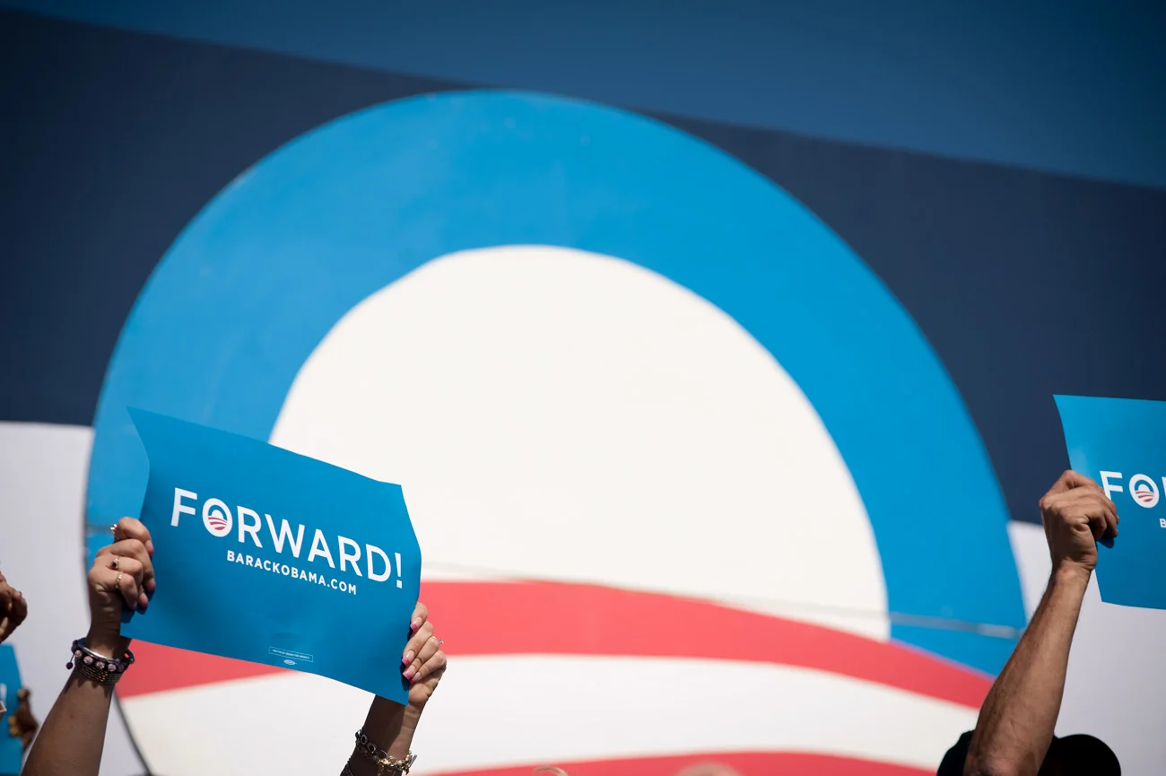 A large rising sun logo in red, white, and blue, rests behind a crowd that is holding up signs that read, “Forward.” The Obama logo is inside the word forward. 