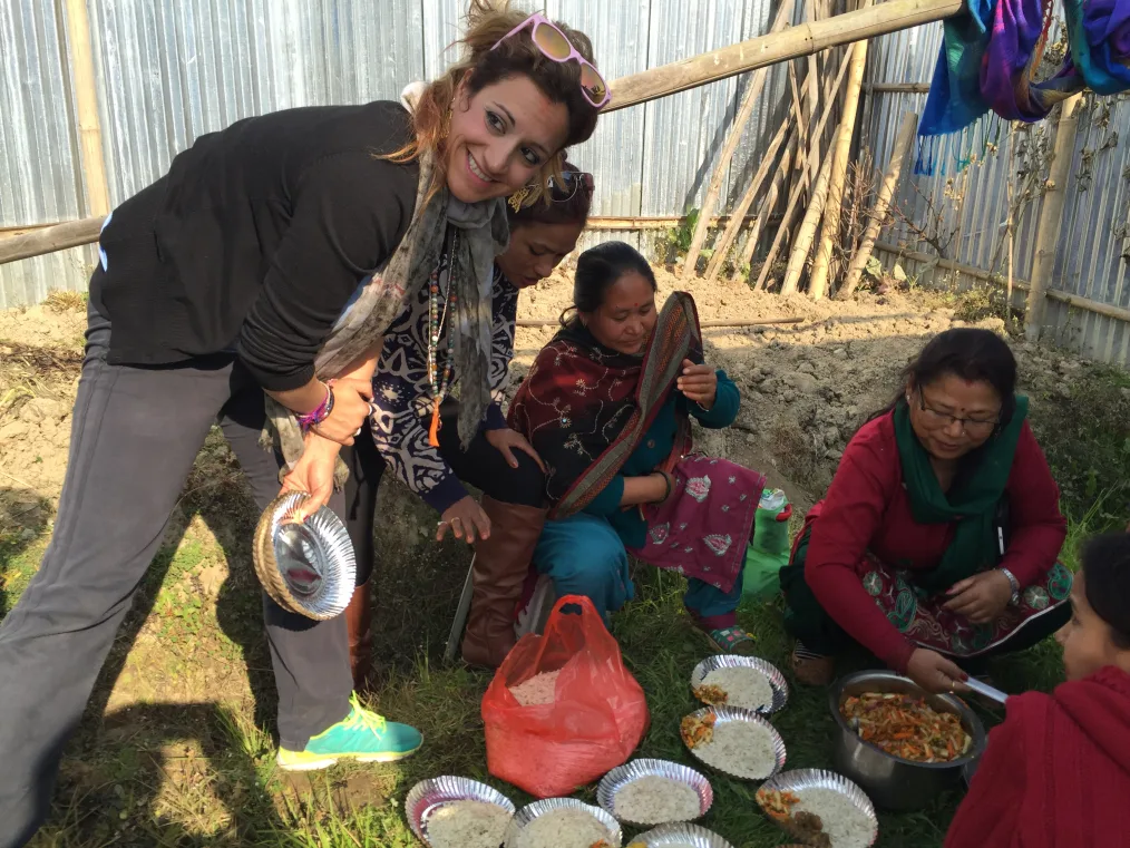 Elpida Kokkota leans next two four women with medium skin tones who are sitting on the ground preparing food. Rice and vegetables are seen on the ground with aluminum pans. 